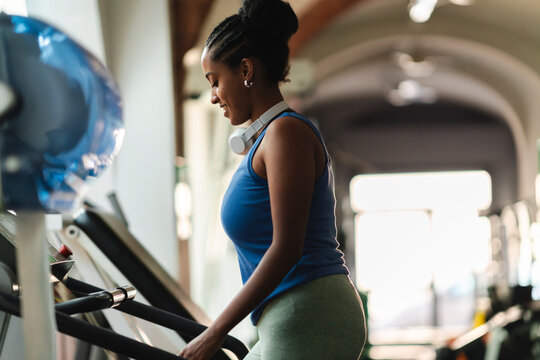 A smiling young Black woman with headphones around her neck exercises on a treadmill in a gym. She looks down, focused on her fitness and healthy lifestyle.
