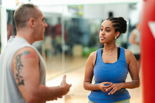 A male personal trainer and his female client are conversing in a gym. He is explaining a workout or fitness concept, and she is attentively listening to his instructions.