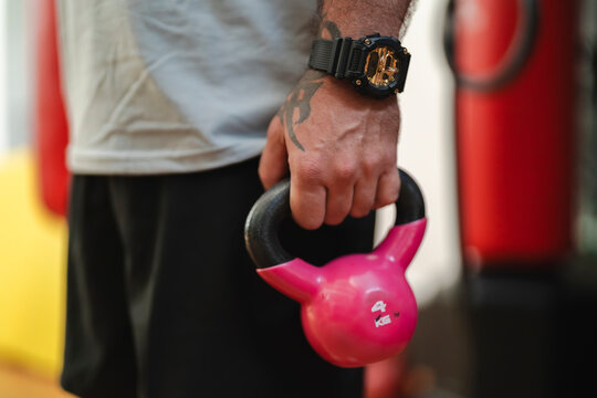 A man holds a pink 4kg kettlebell, showcasing fitness and strength training. His tattooed arm and a black and gold watch are clearly visible in a gym environment.