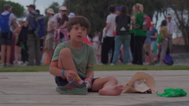 Young boy sitting barefoot on pavement in public square, putting on socks beside scattered belongings, large group of people and activity in background