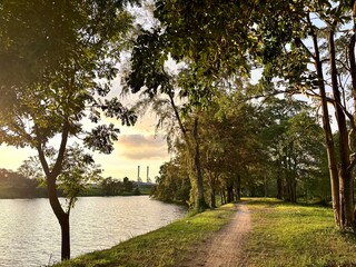 Scenic dirt path beside a tranquil lake with lush trees, silhouetted against a golden sunset sky, with a distant power plant or factory on the horizon