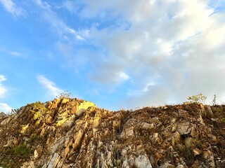 A low-angle shot of a rocky hillside covered in jagged stones and sparse grass, illuminated by the golden light of the late afternoon sun, set against a partly cloudy blue sky.