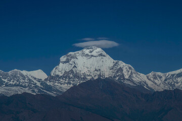 Snow-covered mountain peak beneath a deep blue sky, a lenticular cloud hovering above, with dark rocky foothills in the foreground.