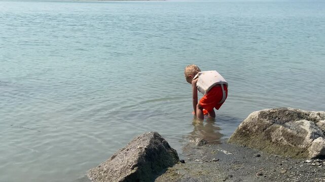 A child in a life vest stands partially in the light blue water at the rocky edge of a lake near Simferopol, Crimea, holding an orange and blue bucket. The moment of focused play and enjoyment