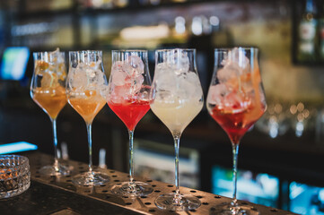 Colorful cocktails with ice cubes in elegant wine glasses lined up on a bar counter, showcasing vibrant drinks in a stylish nightlife setting
