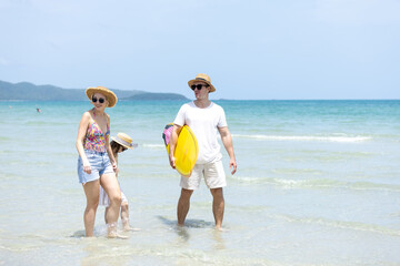 Fototapeta premium Happy family enjoying tropical beach vacation. Father holding yellow surfboard while mother and daughter stand together in shallow water. Concept of summer fun, bonding and seaside lifestyle.