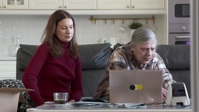 A middle-aged woman is patiently teaching her elderly mother how to use a laptop and thermal label printer for their online store business in a modern, well-lit home setting.