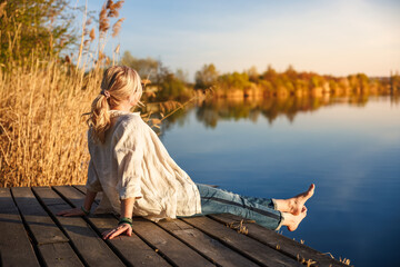 Woman sitting and relaxing barefoot on wooden pier by still lake during golden hour of indian summer