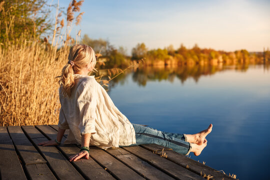 Woman sitting and relaxing barefoot on wooden pier by still lake during golden hour of indian summer - Powered by Adobe