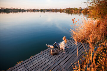 Tranquil lake scenery with woman relaxing on pier while swan swimming in distance. Connection and...