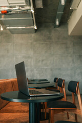 Slim modern laptop on round black table in stylish cafe interior with chairs and concrete wall background. Minimalist workspace concept in urban coffee shop