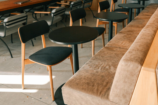 Modern café interior with black round tables, wooden chairs with dark upholstery, long wooden table and minimalist chairs in background, cozy coworking and dining atmosphere - Powered by Adobe