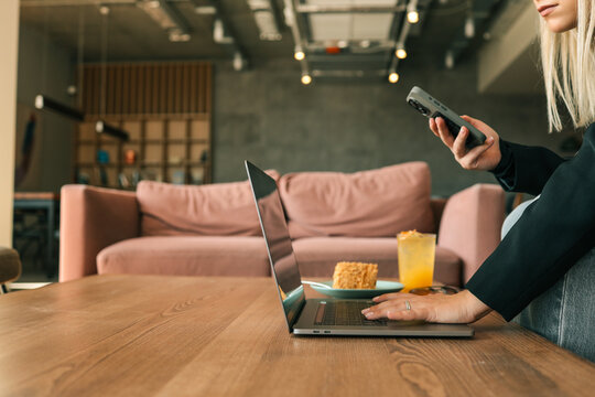 Freelancer woman working on laptop while holding smartphone in cozy cafe. Glass of orange juice and dessert on table, modern interior with soft sofa and stylish casual outfit