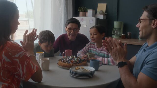 Slowmo shot of middle-aged Caucasian grandmother blowing candles on birthday cake with beloved grandchildren, with young parents clapping hands sharing joyous family moment - Powered by Adobe