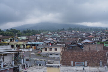 Sunday, August 3, 2025, 14:19:44, Temanggung, Indonesia _ Panoramic view of a densely populated village nestled at the foot of a majestic, cloud-covered mountain.