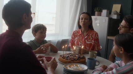 Young beautiful Caucasian mother taking time to make wish before blowing candles on birthday cake, surrounded by happy family members including kids in cozy kitchen, all clapping hands