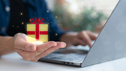 Close-up hand holding floating gift box icon with sparkle, using laptop. Concept of online gift,...
