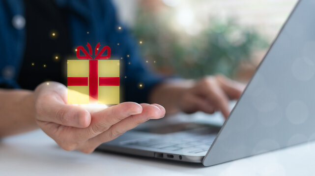 Close-up hand holding floating gift box icon with sparkle, using laptop. Concept of online gift, digital promotion, and virtual reward.