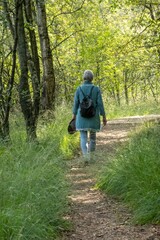 Obraz premium Senior woman walking on path in green forest holding hat and backpack
