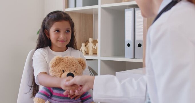 A girl holds a striped teddy while meeting a pediatric doctor for a routine appointment in a bright clinic; they share a friendly handshake to build trust. Girl and doctor share a warm smile.