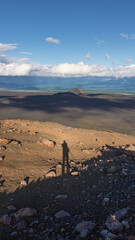 Shadow of a person standing on rocky terrain overlooking a vast landscape with mountains and clouds, capturing the essence of adventure and exploration in nature