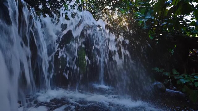 Sunbeams illuminate waterfall cascading jungle rocks, water splashes into stream, forest green surrounds stones. Nature flows gently, waterfall pours into river, green forest wraps water stones.