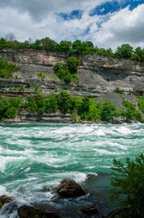 The powerful, turquoise rapids of the Niagara River flowing through a lush green gorge with rocky cliffs. The sky is partly cloudy, and the trees are vibrant green, suggesting a warm and sunny day.