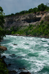 The powerful, turquoise rapids of the Niagara River flowing through a lush green gorge with rocky cliffs. The sky is partly cloudy, and the trees are vibrant green, suggesting a warm and sunny day.