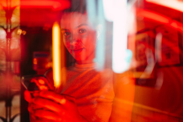 Half-hidden behind neon light, woman looks up from smartphone with faint smile — visual metaphor...