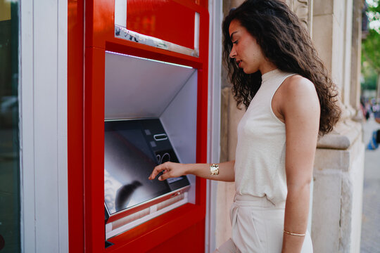 Professional woman at red ATM, engaging with digital banking interface, symbolizing trust, convenience, and adaptability in tech-driven financial services. - Powered by Adobe