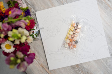 A set of small, numbered acrylic paint pots for a paint-by-numbers kit, sealed in plastic and resting on top of the corresponding canvas with its numbered outlines.