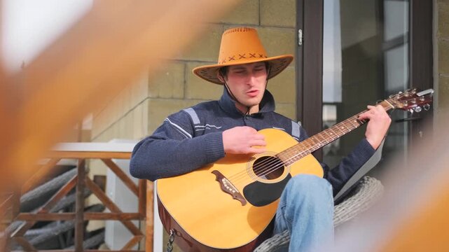Happy man in cowboy hat strumming acoustic guitar outdoors while sitting on a home porch. Rustic background, patterned wood, create a warm, countryside vibe. Music, rural lifestyle, and relaxation.