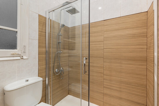 Modern bathroom with a glass shower enclosure and wooden wall panels. White toilet and tiled floor are visible. Bright lighting enhances the clean design.