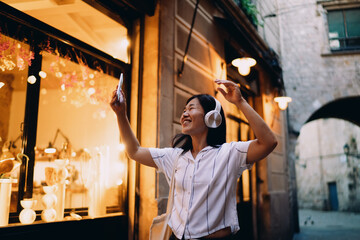 Asian tourist woman raises arms while holding phone, expressing excitement, self-expression and emotional connection through digital technology and mobile joy. Making selfie