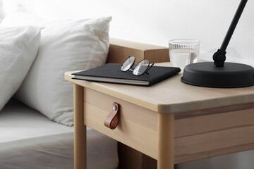 Bedside table with lamp, notebook, glasses and glass of water near bed indoors, closeup