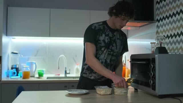 Playful young man tosses a spoon into the air and opens a plastic container with salad, preparing to eat dinner alone in kitchen. Evening routine of an introverted man enjoying a quiet meal at home.