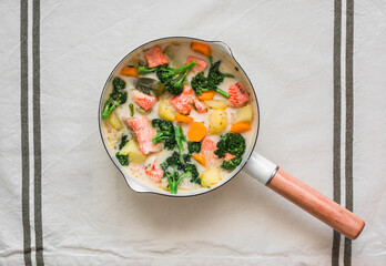 Salmon creamy fish soup with potatoes, carrots and broccoli in a saucepan on a light background, top view