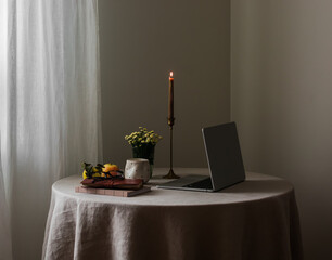 A cozy home interior - a candle in a brass candlestick, a laptop, books, a glass, apples on a round table with a gray linen tablecloth