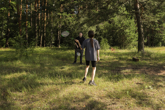 Teen girl preparing to hit shuttlecock while father watches from across the grassy clearing, relaxed atmosphere of family fun and natural wellness.