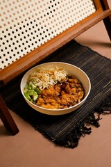 Close three-quarter angle of pempek drenched in cuko alongside springy noodles and crisp cucumber, presented in a ceramic bowl on a textured mat with soft rattan backdrop.