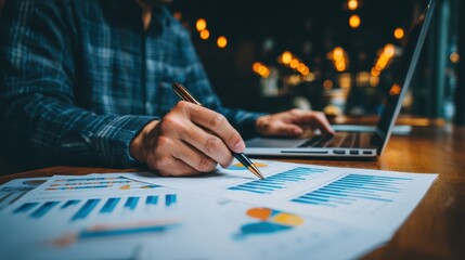 A person reviews financial charts and graphs at a desk with a laptop, analyzing data in a business setting with warm lighting