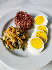 Red rice with vegetable salad or gudangan and a boiled egg on a white plate. A healthy meal concept.