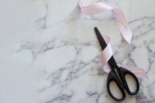 Top view of black scissors cutting pink satin ribbon on marble background copy space