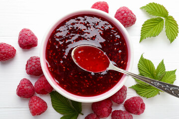 Sweet raspberry jam in bowl, berries and green leaves on white wooden table, flat lay