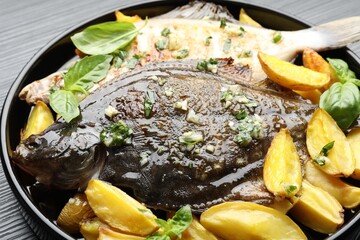Tasty roasted flounder fish with herbs and potato on black table, closeup. Homemade seafood dish