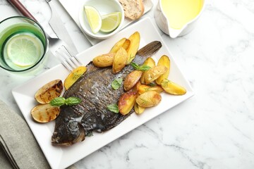 Homemade seafood dish. Tasty roasted flounder fish served with potatoes, basil and lemon slices on white marble table, flat lay. Space for text