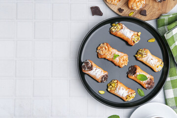 Delicious cannoli with cheese, nuts, powdered sugar, chocolate and mint on white tiled table, flat lay. Space for text