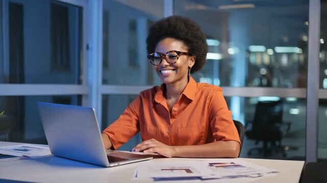 A young professional woman working on her laptop at her desk, late at night. The office setting creates a productive yet intimate atmosphere. The woman is wearing glasses and a bright orange shirt.