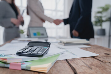 Calculator and documents are lying on a wooden desk in an office, with business people shaking...