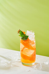 Cold refreshing orange and lime cocktail garnished with mint and thyme leaves in glass on green light background. Summer Drinks for home.